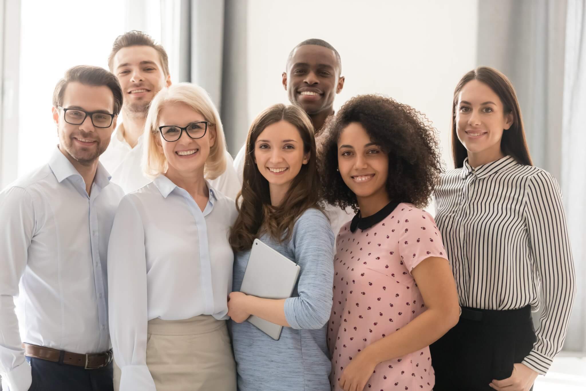 Multiethnic smiling businesspeople standing looking at camera making group photo in office together, happy diverse employees posing for picture with boss or team leader, showing unity and support